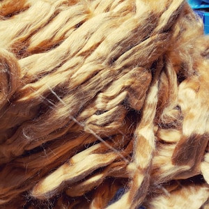 May include: Close-up of a pile of golden-brown fiber, possibly wool or silk. The fibers are soft and textured, with a slightly wavy appearance. The fibers are bundled together, creating a dense, tactile mass.