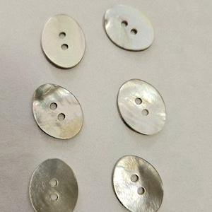 May include: Six oval-shaped, iridescent mother-of-pearl buttons. Each button has two holes for sewing. The buttons have a pearlescent sheen, reflecting light with a subtle iridescence. The buttons are arranged on a light-colored surface.