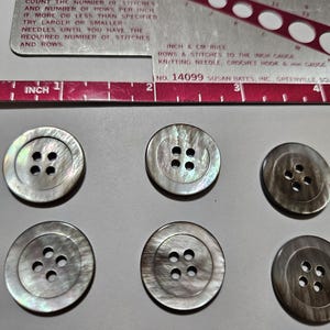 May include: Six iridescent, round buttons with four holes each are arranged on a gray surface. A metal stitch gauge and ruler are in the background, with text indicating inch and cm measurements. The buttons are likely for sewing or crafts.