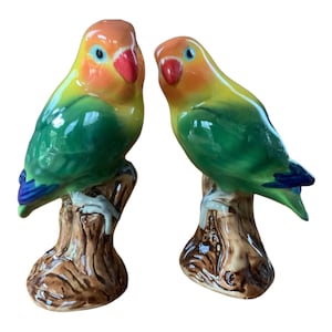 May include: Two ceramic lovebird figurines perched on tree stumps. The birds are green, yellow, and orange with blue accents.
