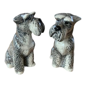Puede incluir: Un par de saleros y pimenteros de cerámica con forma de perros Schnauzer. Los perros están sentados, con detalles de pelaje gris y blanco. Los saleros tienen narices negras y son brillantes.