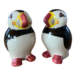 May include: A pair of ceramic salt and pepper shakers shaped like puffins. The puffins are white with black and red accents. The shakers are about 4 inches tall.