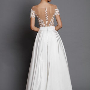Op de afbeelding: Een witte bruidsjurk met een doorschijnend kantbovenstuk en een volle rok. Het bovenstuk is versierd met delicate bloemenborduursels.