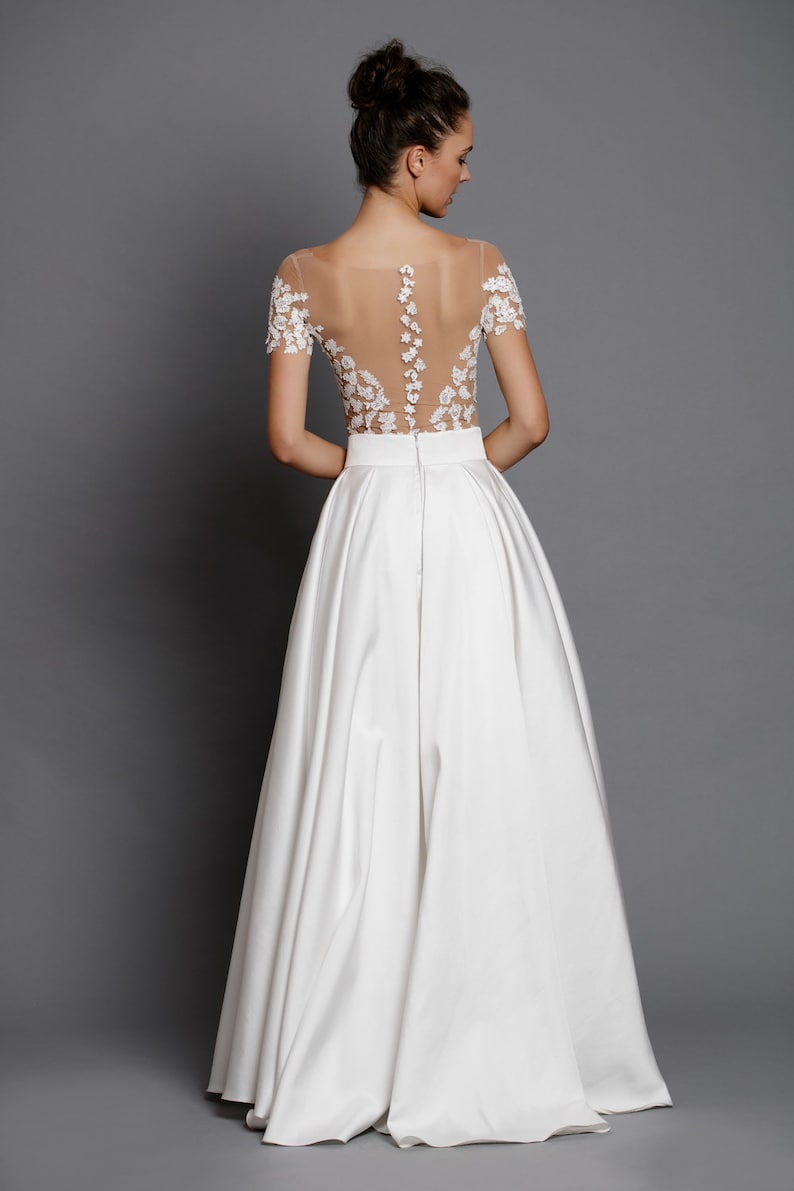 Op de afbeelding: Een witte bruidsjurk met een doorschijnend kantbovenstuk en een volle rok. Het bovenstuk is versierd met delicate bloemenborduursels.