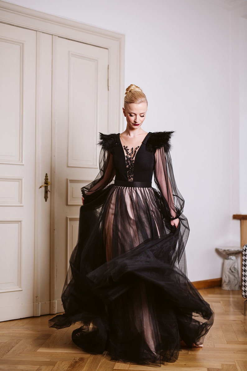 May include: A woman in a black and pink tulle gown with a black bodice and lace detailing. The gown has long sleeves with feathered accents on the shoulders. She is standing in a room with a white door in the background.