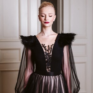 May include: A woman wearing a black gown with sheer sleeves and a black feather trim. The gown has a plunging neckline with a black lace overlay. The woman has her eyes closed and is standing in front of a white door.