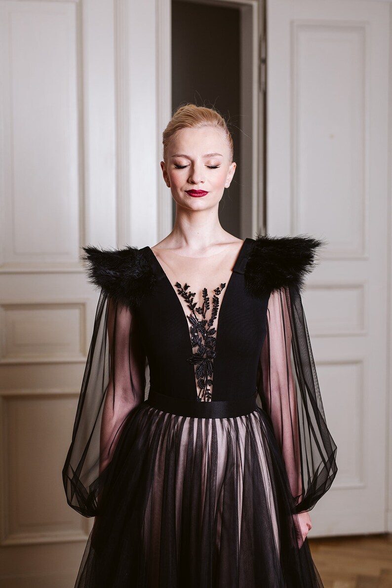 May include: A woman wearing a black gown with sheer sleeves and a black feather trim. The gown has a plunging neckline with a black lace overlay. The woman has her eyes closed and is standing in front of a white door.