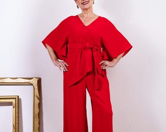 May include: A woman wearing a red jumpsuit with a wide-leg silhouette. The jumpsuit has a wrap-style top with a self-tie belt. The woman is smiling and looking to the side.