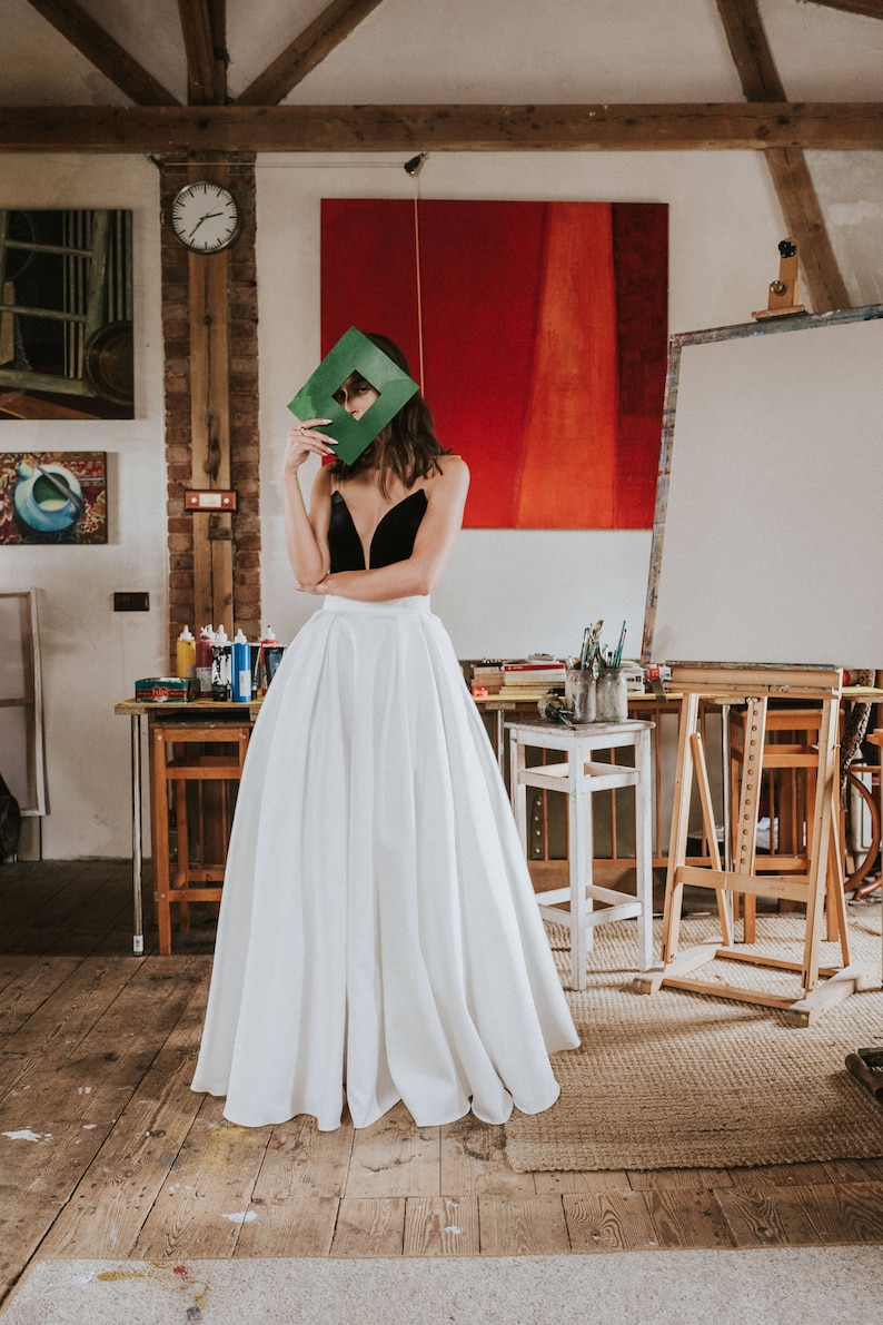 Op de afbeelding: Een vrouw in een zwart fluwelen lijfje en een witte lange rok staat in een kunststudio. Ze houdt een groen vierkant voor haar gezicht. De studio heeft een houten vloer, een houten ezel en een rood en oranje abstract schilderij aan de muur.