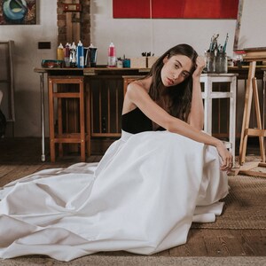 Op de afbeelding: Een vrouw in een witte jurk met een zwart lijfje zit in een kunststudio. De jurk is vloeiend en golft om haar heen. De studio is gevuld met kunstbenodigdheden en ezel.
