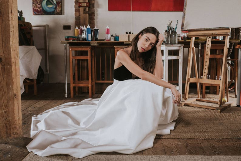 Op de afbeelding: Een vrouw in een witte jurk met een zwart lijfje zit in een kunststudio. De jurk is vloeiend en golft om haar heen. De studio is gevuld met kunstbenodigdheden en ezel.