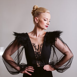 May include: A woman wearing a black dress with sheer black sleeves and feather detailing on the shoulders. The dress has a plunging neckline and a lace overlay.