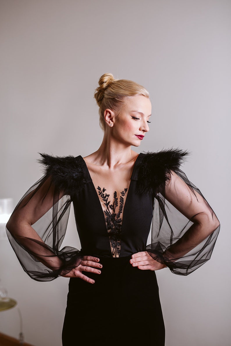 May include: A woman wearing a black dress with sheer black sleeves and feather detailing on the shoulders. The dress has a plunging neckline and a lace overlay.