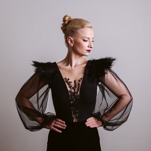 May include: A woman wearing a black dress with sheer sleeves and feather detailing on the shoulders. The dress has a deep V-neckline and a lace overlay.