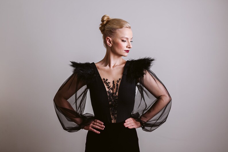 May include: A woman wearing a black dress with sheer sleeves and feather detailing on the shoulders. The dress has a deep V-neckline and a lace overlay.