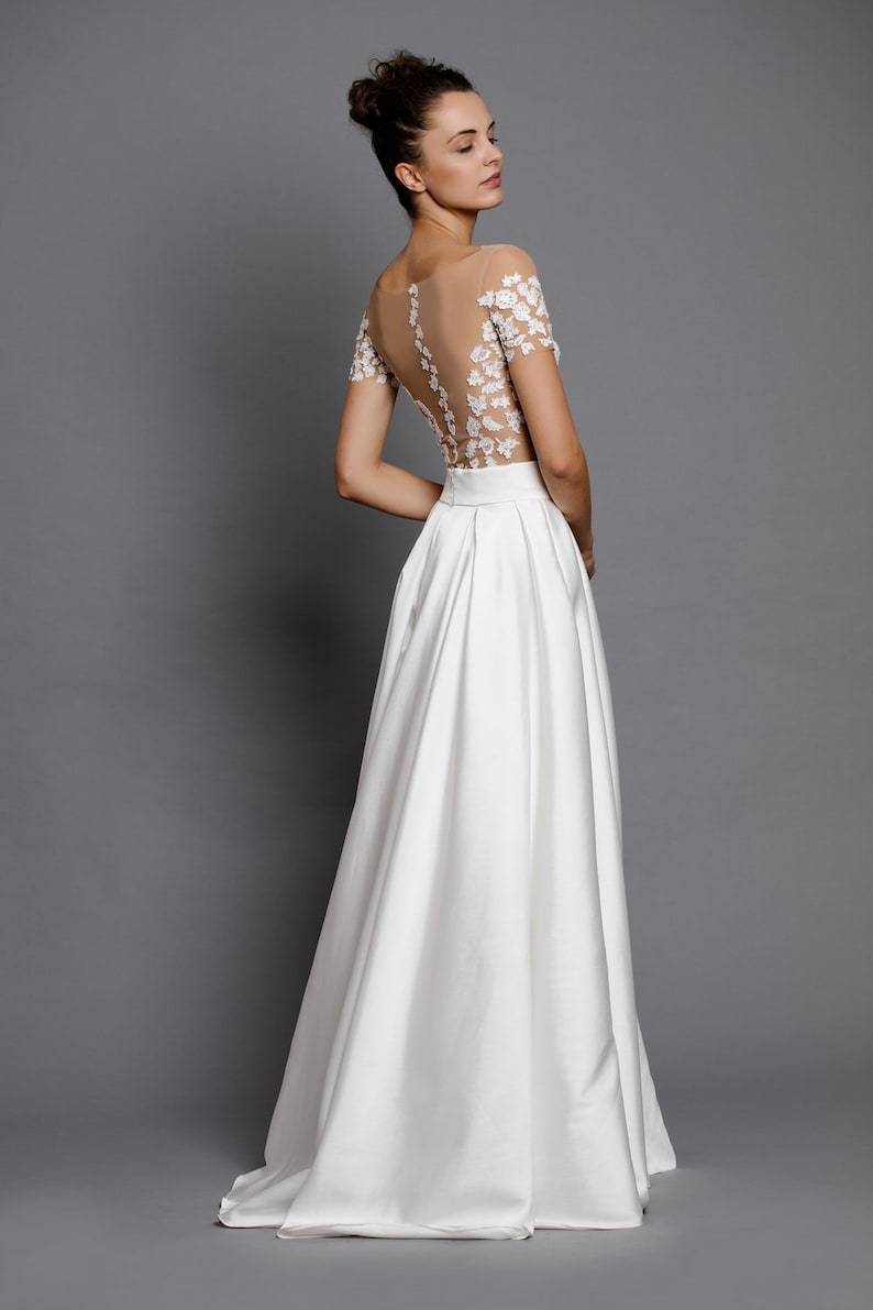 Op de afbeelding: Een witte bruidsjurk met een doorschijnend kantbovenstuk en een volle rok. Het bovenstuk is versierd met delicate bloemenborduursels.