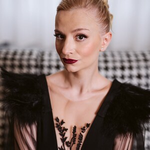 May include: A woman wearing a black sheer dress with feather detailing on the shoulders and a black floral lace overlay. She has her blonde hair pulled back in a bun and is wearing red lipstick.
