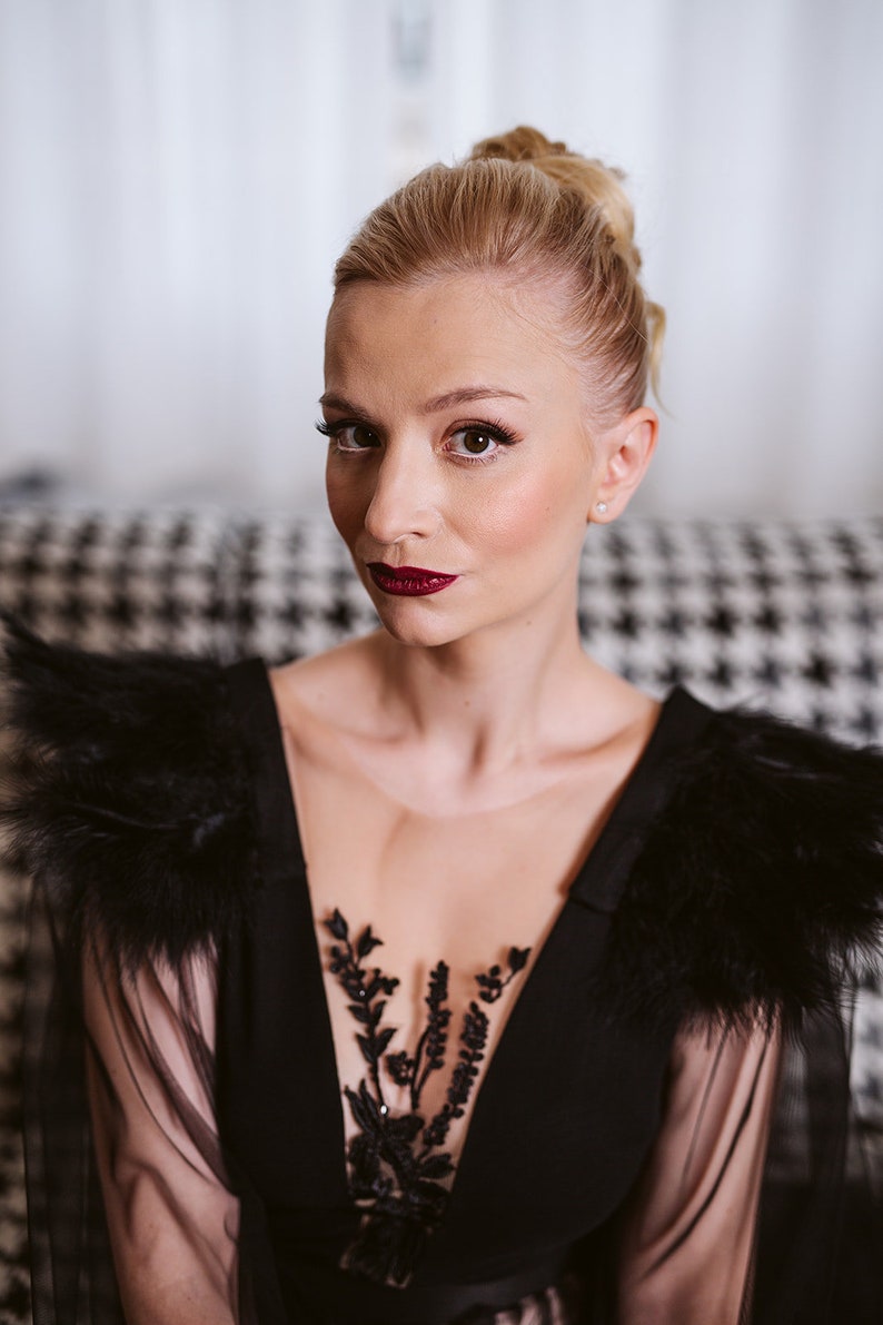 May include: A woman wearing a black sheer dress with feather detailing on the shoulders and a black floral lace overlay. She has her blonde hair pulled back in a bun and is wearing red lipstick.