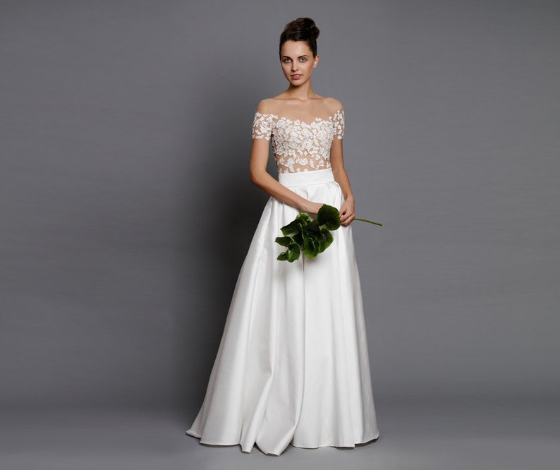 Op de afbeelding: Een witte, off-shoulder bruidsjurk met een kantbovenstuk en een zwierige rok. Het model houdt een enkel groen blad vast.