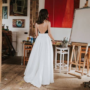 Op de afbeelding: Een vrouw draagt een witte maxi-rok en een zwart, mouwloos topje met een lage rug. Ze staat in een kunststudio met een rood schilderij op een ezel achter haar.