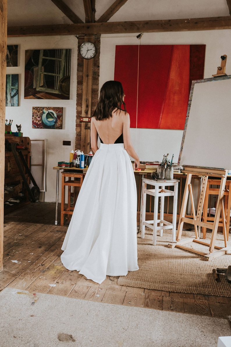 Op de afbeelding: Een vrouw draagt een witte maxi-rok en een zwart, mouwloos topje met een lage rug. Ze staat in een kunststudio met een rood schilderij op een ezel achter haar.