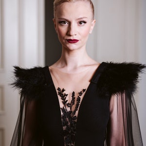 May include: A woman wearing a black gown with a sheer overlay and feather detailing on the shoulders. The gown has a plunging neckline and a floral lace design on the bodice.