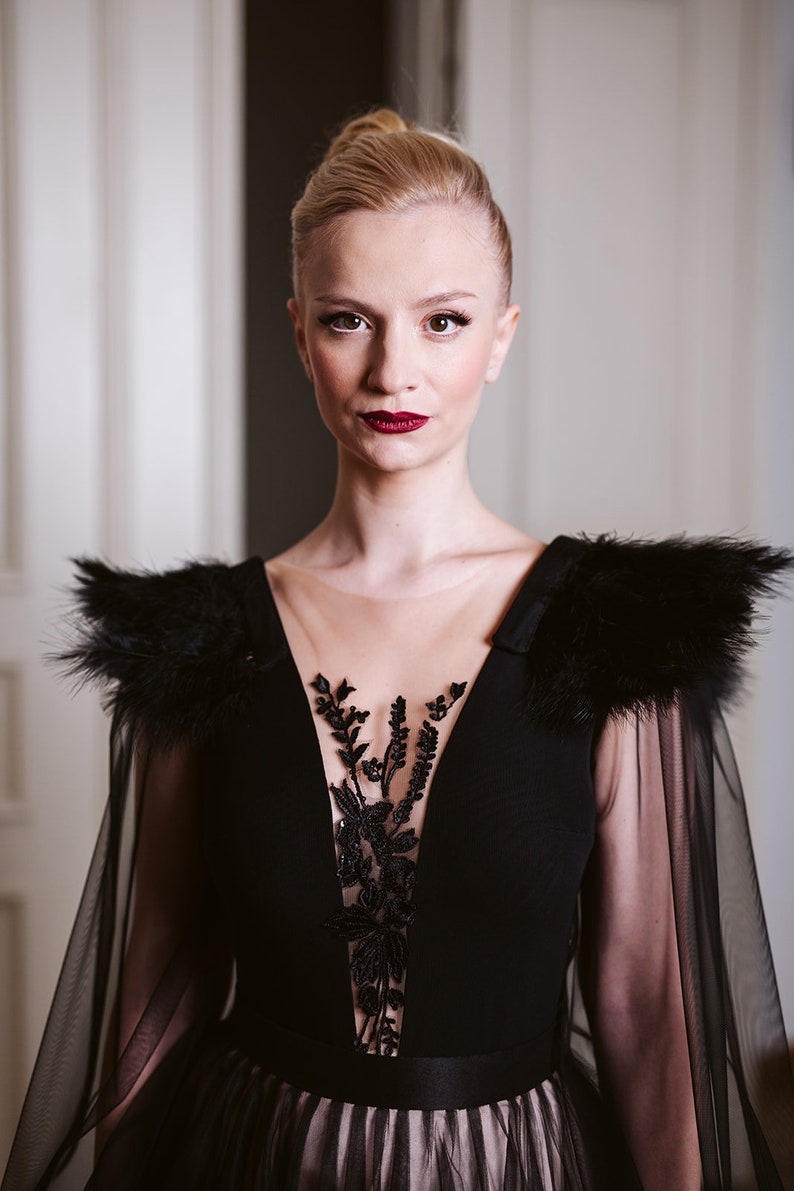 May include: A woman wearing a black gown with a sheer overlay and feather detailing on the shoulders. The gown has a plunging neckline and a floral lace design on the bodice.