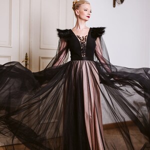 May include: A woman wearing a black gown with a sheer overlay and feathered shoulders. The gown has a black bodice with a lace design and a pink skirt.