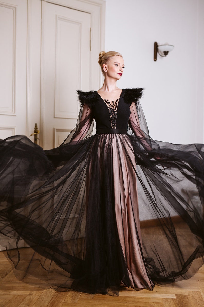 May include: A woman wearing a black gown with a sheer overlay and feathered shoulders. The gown has a black bodice with a lace design and a pink skirt.