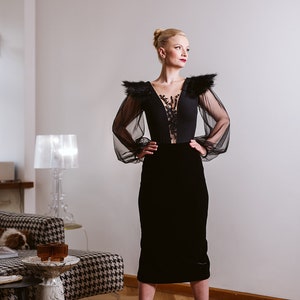 May include: A woman wearing a black velvet skirt and a black sheer top with lace detailing and feather accents. She is standing in a room with a white wall and a lamp in the background.