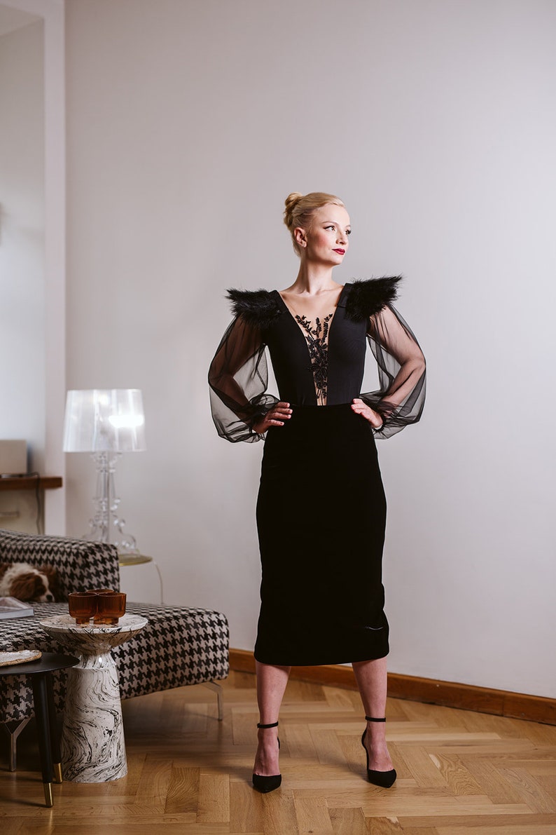 May include: A woman wearing a black velvet skirt and a black sheer top with lace detailing and feather accents. She is standing in a room with a white wall and a lamp in the background.