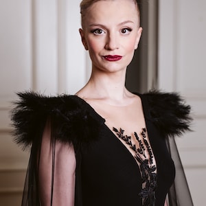 May include: A woman wearing a black gown with a sheer overlay and feather detailing on the shoulders. The gown has a plunging neckline and a black lace design on the bodice. The woman has blonde hair styled in a bun and is wearing red lipstick.