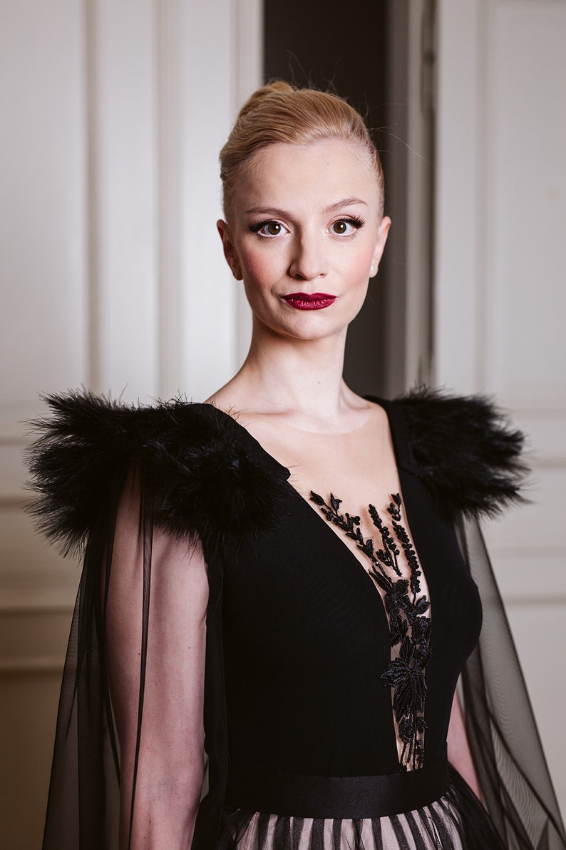 May include: A woman wearing a black gown with a sheer overlay and feather detailing on the shoulders. The gown has a plunging neckline and a black lace design on the bodice. The woman has blonde hair styled in a bun and is wearing red lipstick.
