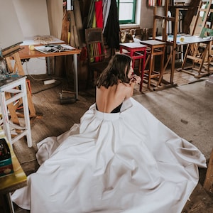 Op de afbeelding: Een vrouw in een witte, vloeiende jurk met een zwarte bustier zit op een houten vloer in een studio. De jurk is rond haar benen gebundeld en de stof is glad en zijdeachtig.