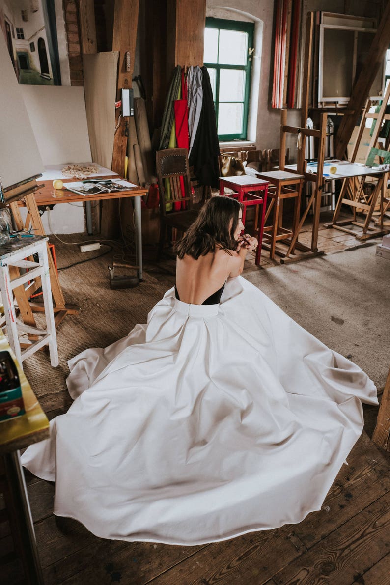 Op de afbeelding: Een vrouw in een witte, vloeiende jurk met een zwarte bustier zit op een houten vloer in een studio. De jurk is rond haar benen gebundeld en de stof is glad en zijdeachtig.