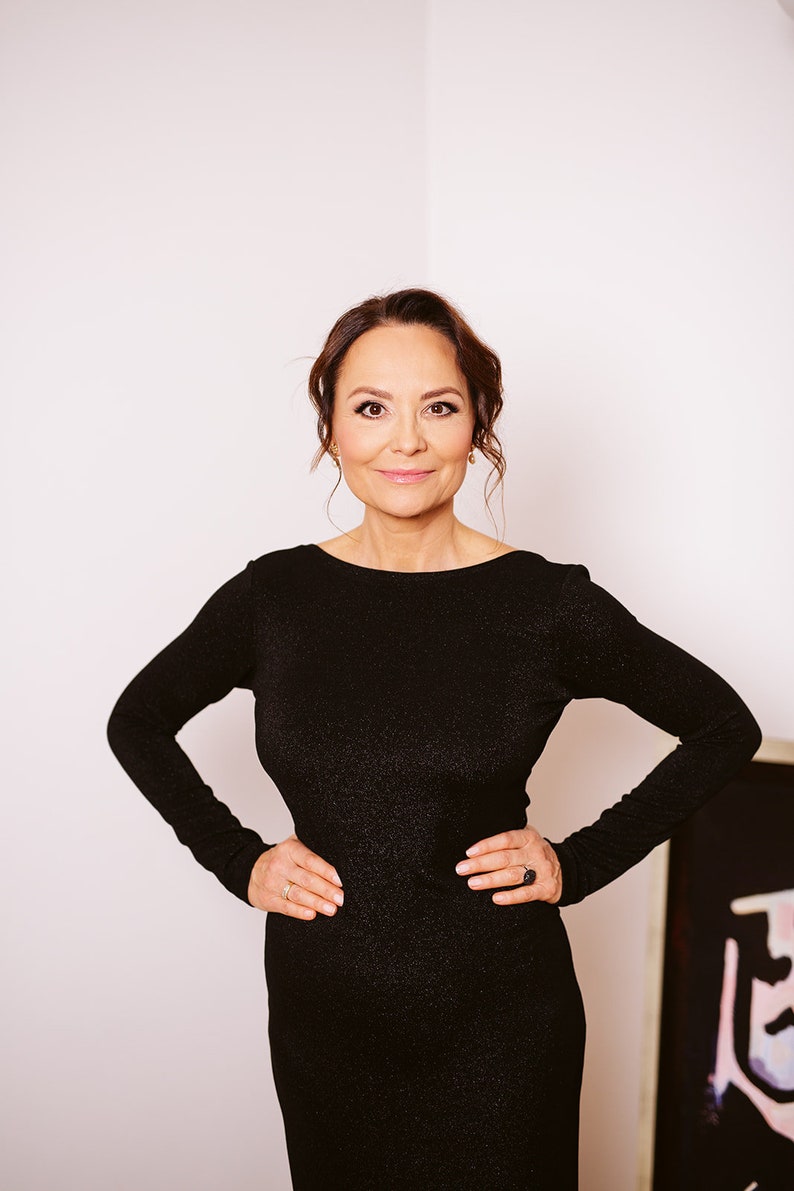 Puede incluir: Una mujer con cabello casta&ntilde;o oscuro lleva un vestido negro brillante. Est&aacute; de pie con las manos en las caderas y mirando directamente a la c&aacute;mara.