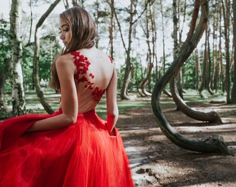 May include: A woman in a red tulle dress with lace detailing stands in a forest of twisted trees. The dress has a sheer back and a fitted bodice.
