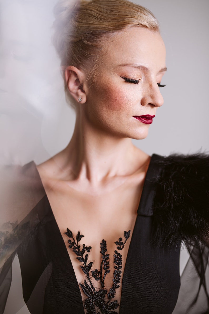 May include: A woman with blonde hair styled in a bun wears a black dress with a sheer lace bodice and a black fur stole. The dress features a floral design on the bodice.