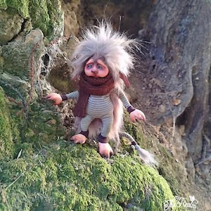 Może przedstawiać: Kapryśna, ręcznie robiona lalka z szarymi, kolczastymi włosami, koszulą w paski i brązowym szalikiem. Lalka ma smutny wyraz twarzy i jest ustawiona na mszystej powierzchni. Lalka ma ogon i nosi spodnie w paski. To fantastyczne stworzenie.