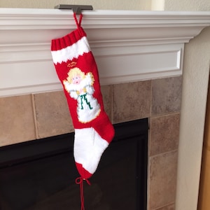May include: A red and white knitted Christmas stocking with a white angel design. The stocking is hanging from a metal hook on a white mantel.