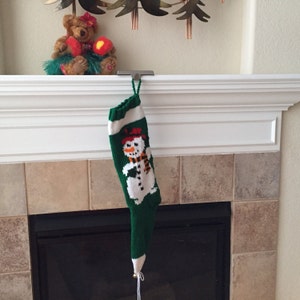 May include: A green knitted Christmas stocking with a snowman design hangs from a metal hook on a white mantel above a fireplace.