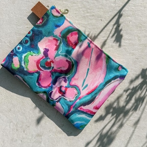 May include: A small, square pouch featuring a colourful floral design in pink, teal, and blue hues. It has a brown leather pull tab and a gold zip. The pattern resembles an abstract watercolour painting.