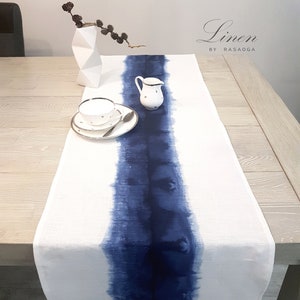 Puede incluir: Un camino de mesa de lino blanco con una franja azul de acuarela en el centro. El texto "Linen by Rasaoga" está impreso en negro en la parte superior del camino de mesa.