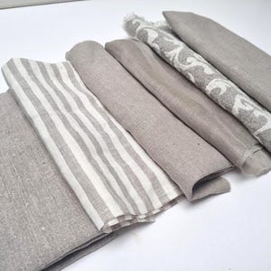 Puede incluir: Colección de servilletas de lino dobladas en varios tonos de gris y beige. Algunas servilletas presentan un estampado de rayas, mientras que otras tienen un color sólido o un diseño decorativo blanco y gris. Las servilletas están dispuestas sobre una superficie blanca.