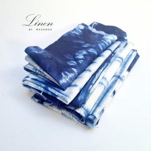 May include: A stack of blue and white tie-dye linen napkins. The napkins are folded and stacked on a white surface. The text "Linen by Rasaoga" is visible at the top of the image.
