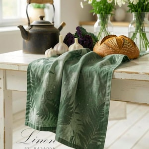 Puede incluir: Un paño de cocina verde con un patrón de hojas de helecho blancas sobre una mesa blanca. El texto Linen BY RASAOGA aparece en la esquina inferior izquierda. La escena incluye una tetera de metal oscuro, bulbos de ajo y una hogaza de pan.
