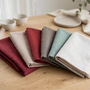 May include: A set of six folded fabric napkins in various colours including deep red, beige, dark grey, sage green, and off-white. Each napkin features a square shape with a visible frayed edge detail along one side, stacked neatly in a row.