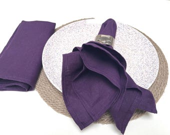 May include: A folded purple linen napkin sits on a white speckled plate, held by a silver napkin ring. Another folded napkin rests to the left. The plate is on a woven placemat, with a white background.