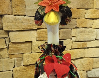 May include: A white goose figurine wearing a black and green dress with a red poinsettia and a red gift box. The dress has a white lace trim and a pattern of small bears and snowflakes. The goose is wearing a black and green hat with a red poinsettia.