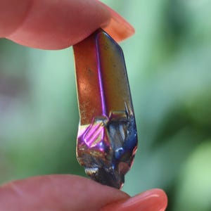 May include: A small, iridescent crystal with a polished, rectangular shape. The crystal displays a spectrum of colors, including gold, purple, and blue, with a rough, textured base. The crystal is held between two fingers against a blurred green background.